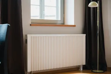 Biały grzejnik pod oknem, między ciemnymi zasłonami, obok stojącej lampy podłogowej.