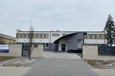 Nowoczesny budynek szkoły za ogrodzeniem, z wejściem i wybrukowanym podjazdem.