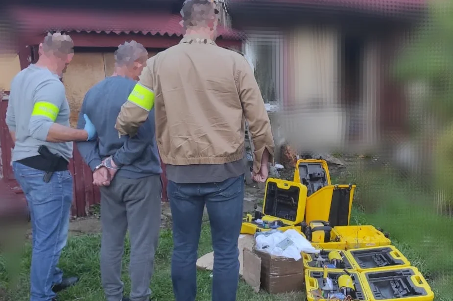 Funkcjonariusze z żółtymi opaskami zatrzymują mężczyznę przed domem, na trawie leżą otwarte żółte walizki ze sprzętem.