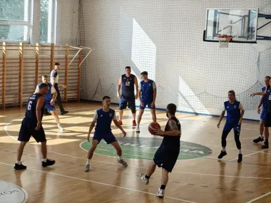 Grupa osób gra w koszykówkę w hali sportowej z drewnianą podłogą, siatką na ścianie i koszem do koszykówki.