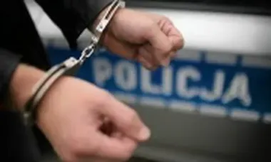 Skute kajdankami dłonie mężczyzny na tle policyjnego pojazdu z napisem POLICJA.