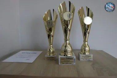 Trzy złote puchary sportowe za pierwsze, drugie i trzecie miejsce oraz dyplom leżą na drewnianym blacie.