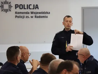 Policjant w mundurze prezentuje dokument grupie funkcjonariuszy podczas spotkania w sali Komendy Wojewódzkiej Policji z siedzibą w Radomiu.