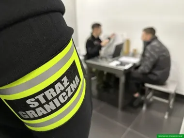 Funkcjonariusz Straży Granicznej w pomieszczeniu służbowym podczas czynności z mężczyzną siedzącym przy biurku.