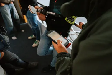 Pasażerowie w środku komunikacji miejskiej, jedna osoba trzyma smartfon, inna osoba z odblaskową opaską na rękawie sprawdza dokumenty.