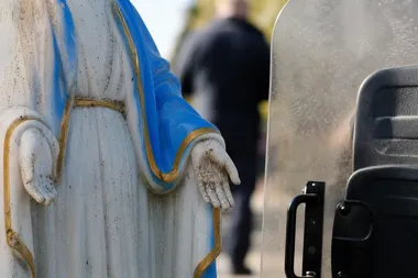 Fragment białej figury z błękitnym szatą stoi obok policyjnej tarczy ochronnej, w tle rozmazana postać mężczyzny.
