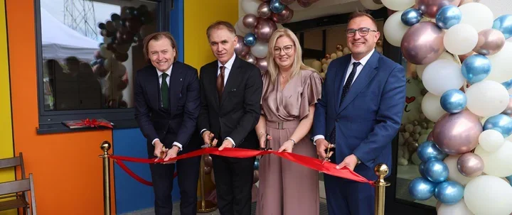 Cztery osoby w eleganckich strojach przecinają czerwoną wstęgę podczas uroczystego otwarcia budynku ozdobionego kolorowymi balonami.